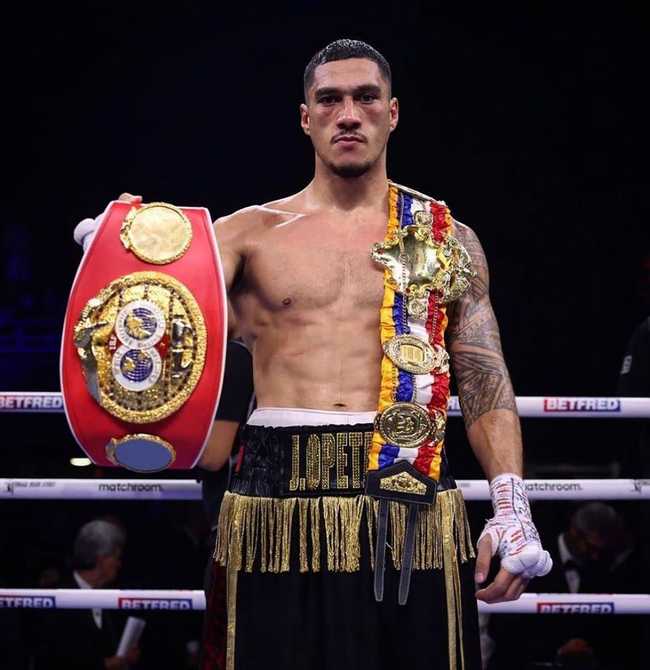Jai Opetiai standing in the ring after a fight holding the red IBF belt in his right hand with the ring belt over his left shoulder