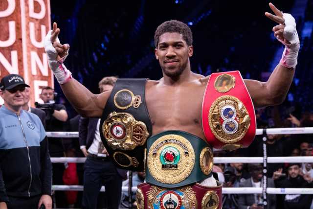 Anthony Joshua in a boxing ring wearing his belts with his arms in the air