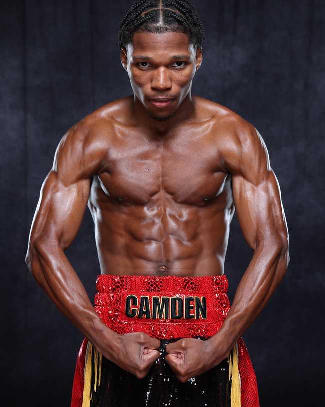 Raymond Ford looking directly at the camera, flexing his muscles and wearing red and black shorts with yellow trim. The shorts have Camden written on the waistband.