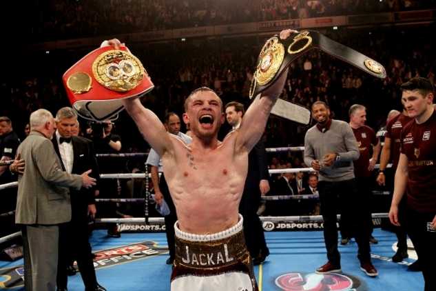 Carl Frampton in a boxing ring holding two belts aloft in victory